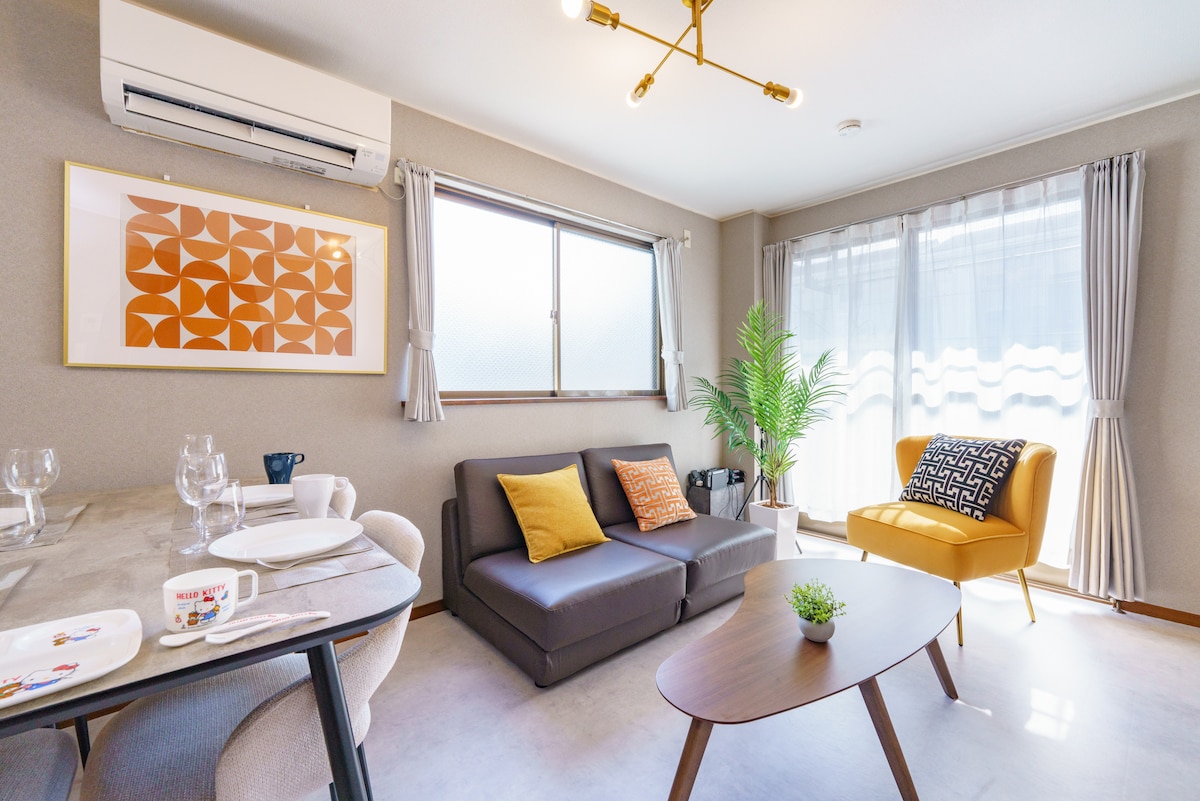 A comfortable living space features a stylish gray sofa paired with a vibrant yellow armchair. A modern dining table is set for meals, while natural light filters through sheer curtains at the windows. Decorative plants and artwork add visual interest to the inviting atmosphere.