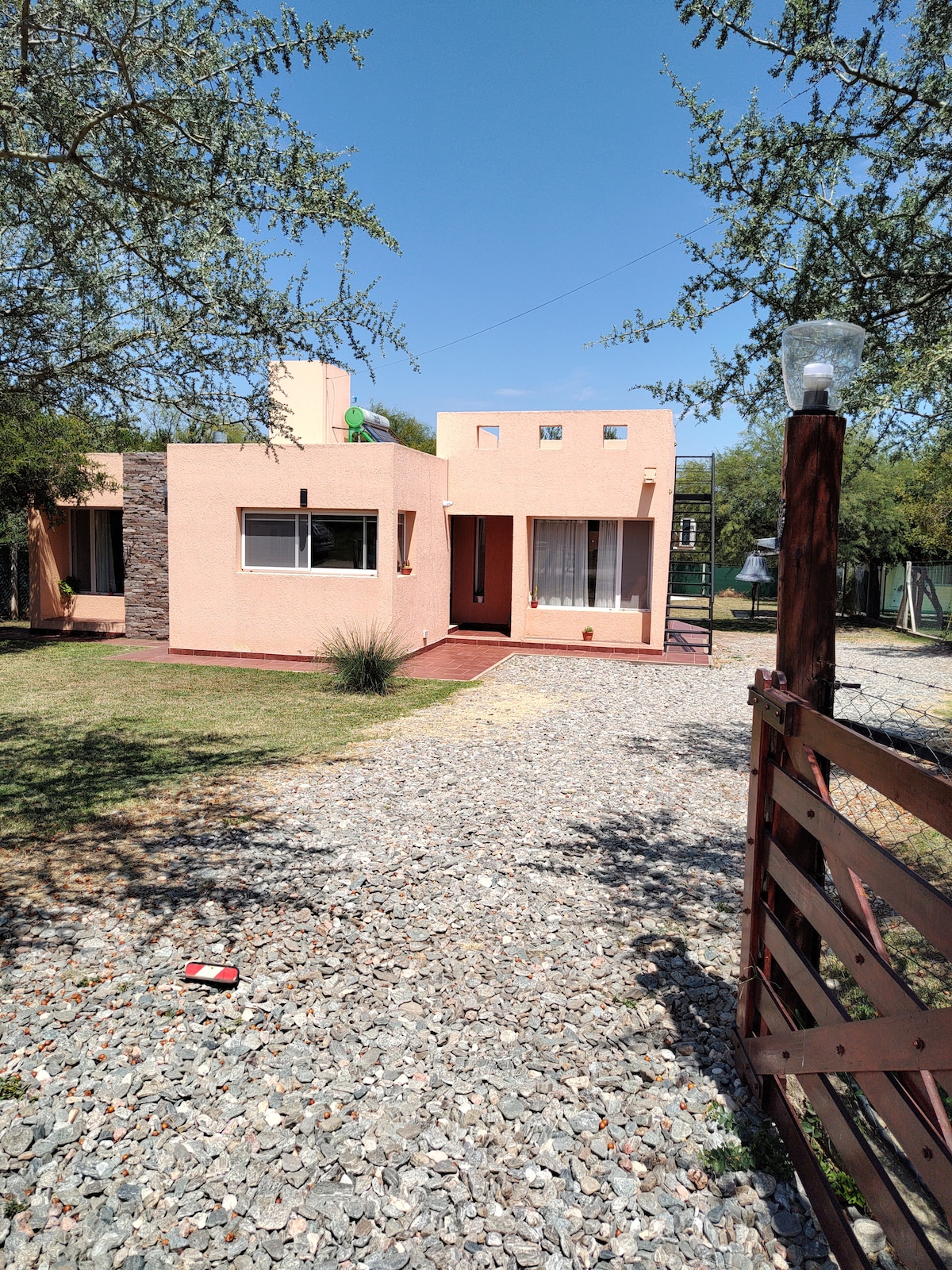 A single-story house is set amidst a gravel pathway and grassy areas, surrounded by trees. The exterior features a blend of stone and soft pastel walls, with large windows allowing natural light to enter. A welcoming front porch is visible, enhancing the outdoor experience.