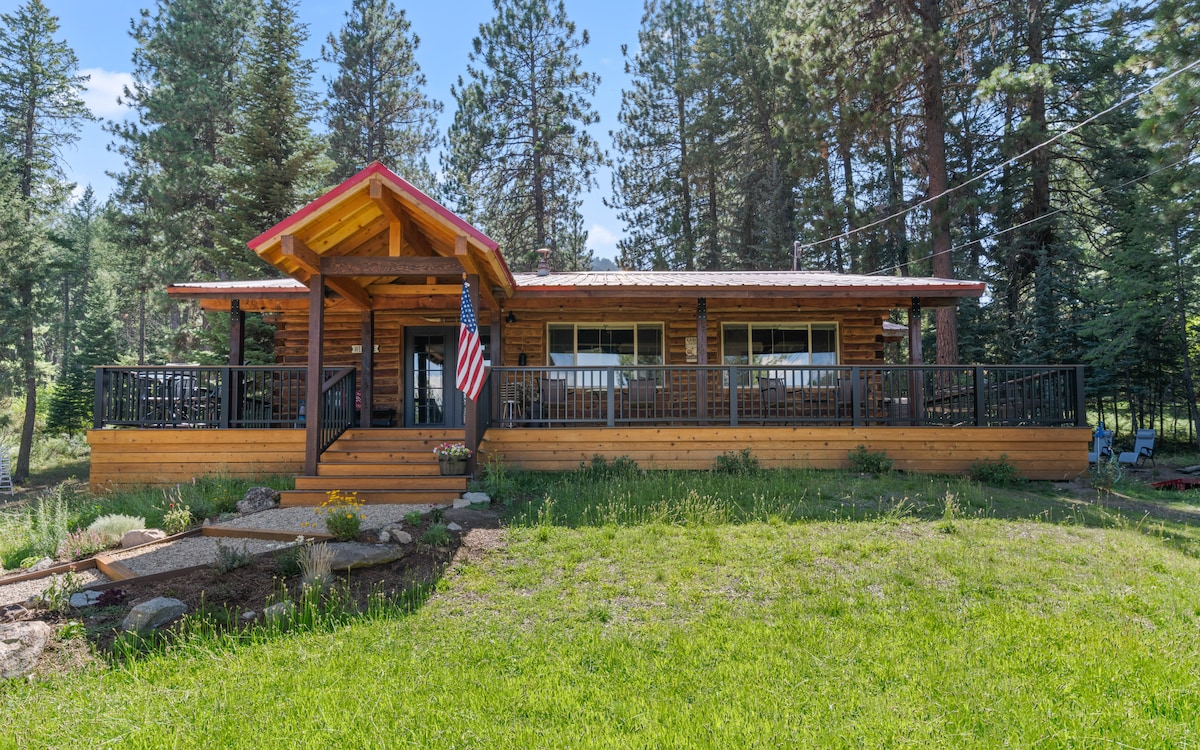 The cabin, constructed from natural logs, features a welcoming porch with a red roof. A wooden deck extends across the front, providing access to the scenic surroundings marked by green grass and towering pine trees.