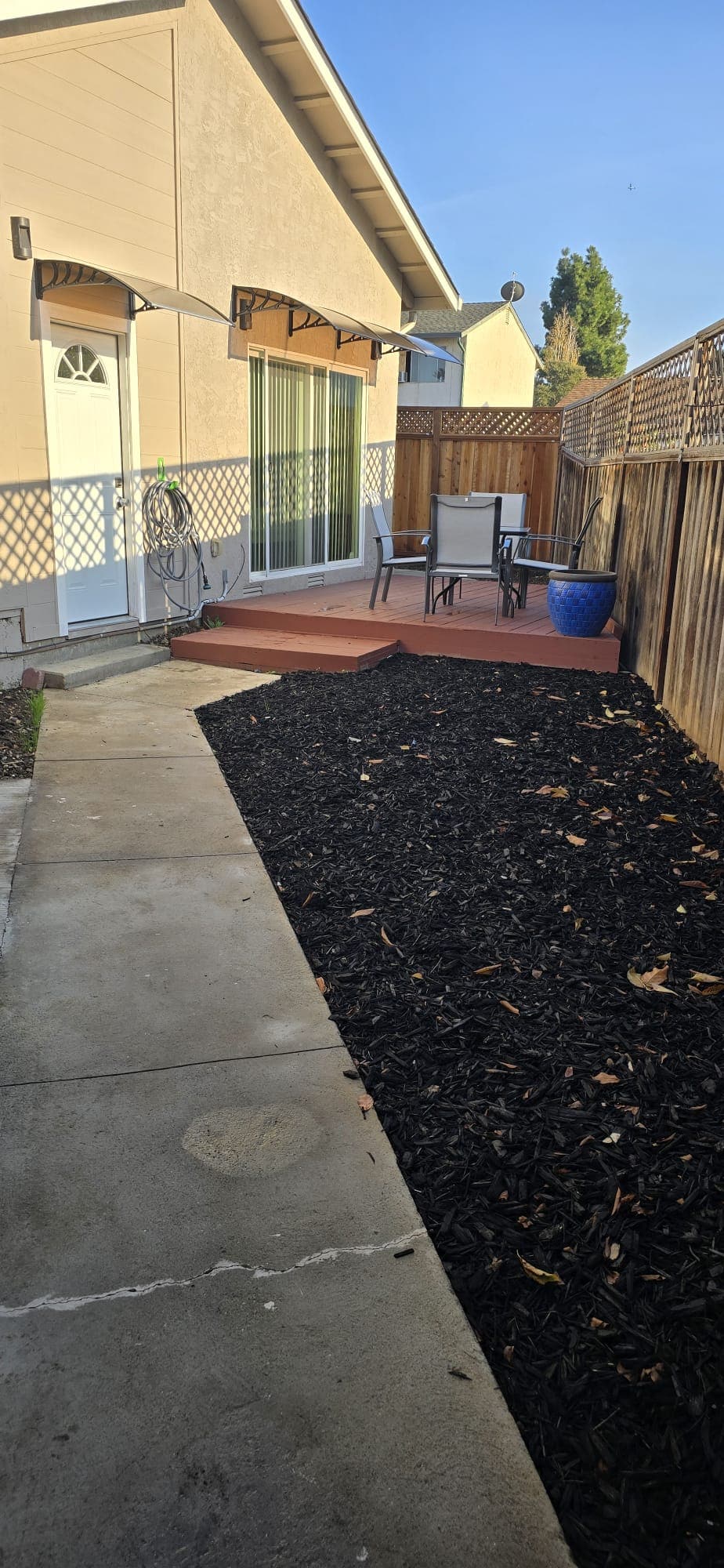 A private patio area is showcased, featuring a redwood deck with two chairs and a small table. Surrounding the patio, dark mulch covers the ground, providing a contrast to the light-colored walls of the house. Natural light enhances the inviting ambiance.