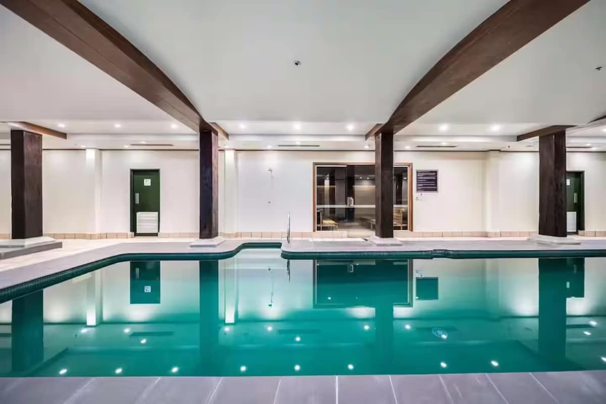 An indoor swimming pool features a tranquil turquoise hue, surrounded by sleek tiles and illuminated by overhead lights. Wooden beams extend across the ceiling, enhancing the modern design. Lounge chairs and a view of the sauna area are visible in the background.