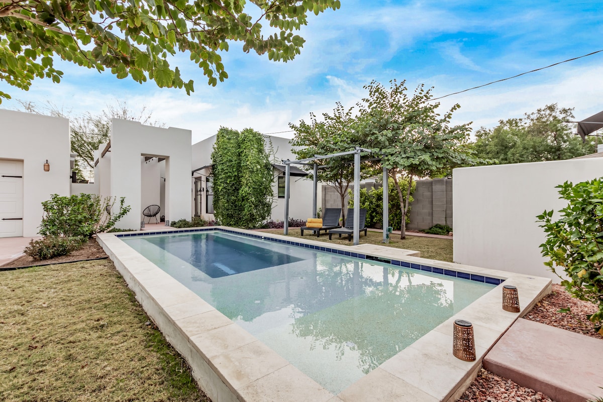 A resort-style pool is situated in a landscaped yard, surrounded by lush greenery and shaded seating. The pool features a clean, reflective surface and a light stone deck, blending seamlessly with the tranquil outdoor environment.