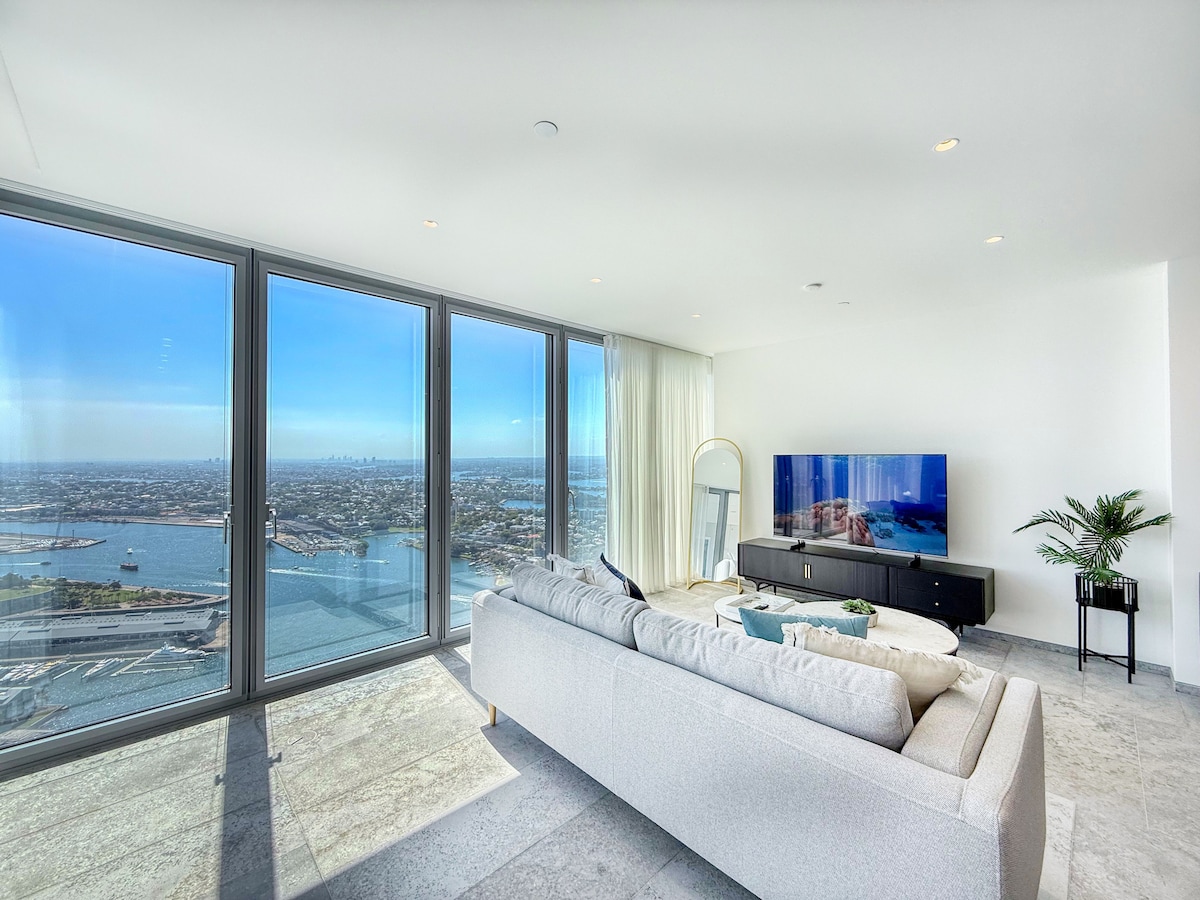 A spacious living area showcases large floor-to-ceiling windows, offering expansive views of Sydney Harbour. A comfortable modular sofa is positioned for relaxation, with a sleek TV stand beneath a mounted television. Natural light fills the space, enhancing the modern design.