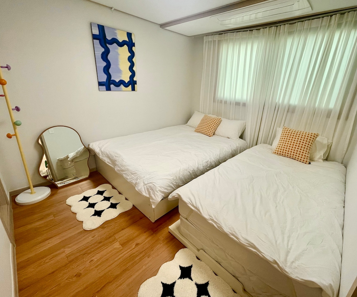 The bedroom features two beds with light-colored bedding, complemented by decorative pillows. A large mirror stands beside a colorful wall art piece, while the floor is adorned with textured area rugs. Sheer curtains allow natural light to filter softly into the space.