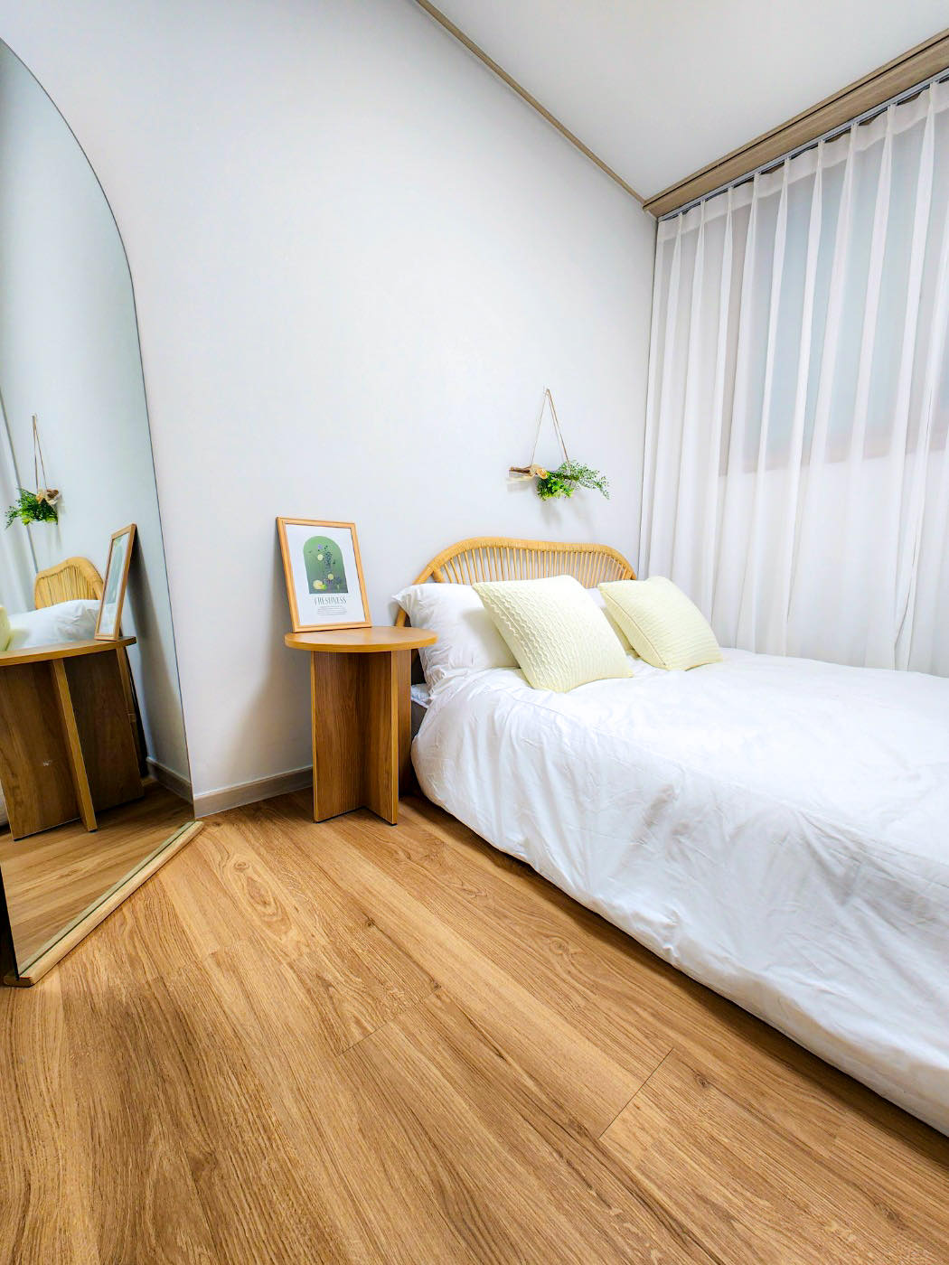 A cozy bedroom features a spacious bed with simple white bedding and two soft pillows. Natural light filters through sheer curtains, creating a calm atmosphere. A small wooden bedside table holds a framed artwork, and a mirror reflects the serene decor. Warm wooden flooring completes the inviting look.