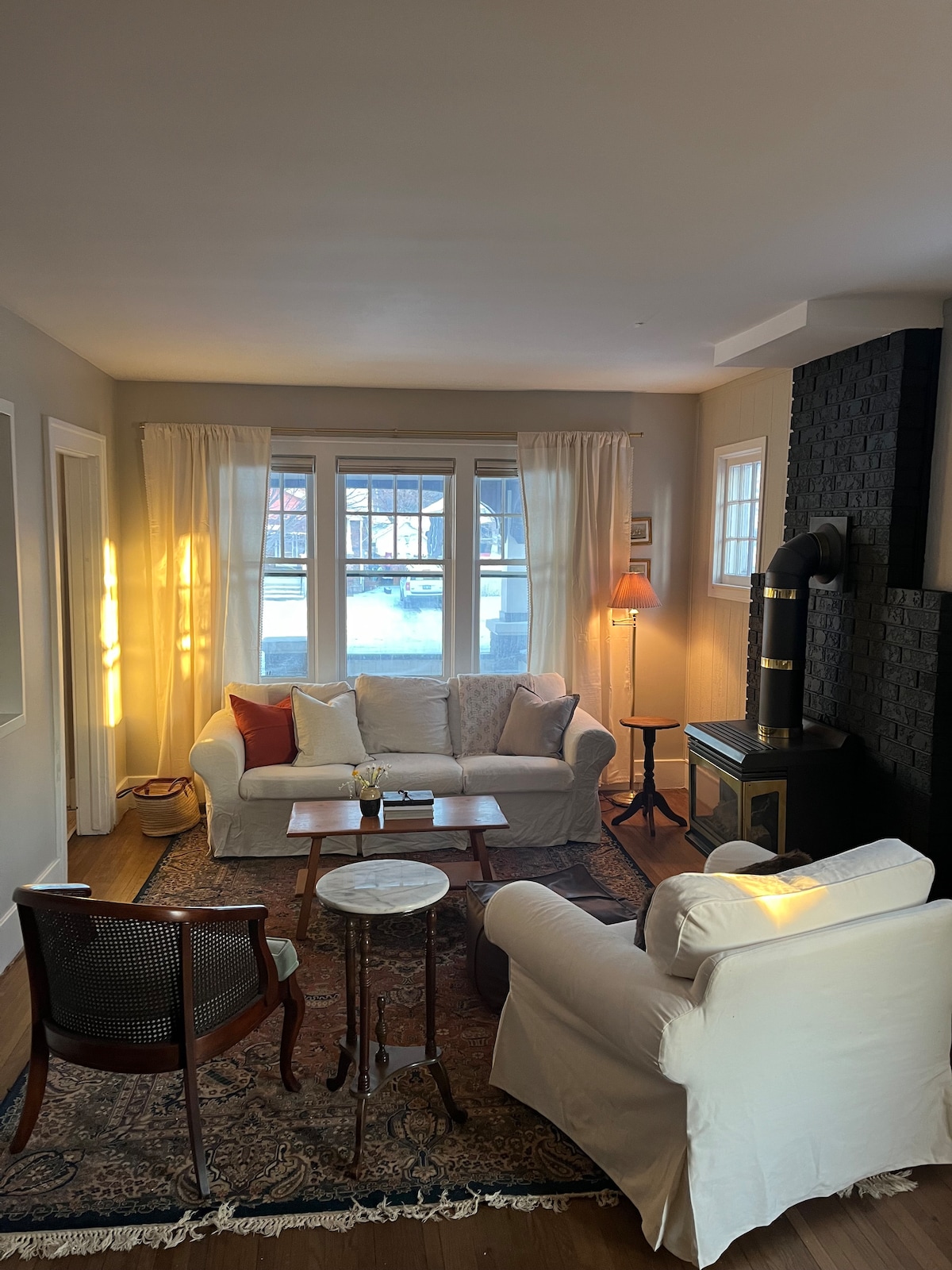 The living area features a comfortable seating arrangement with two white sofas and a wooden chair, arranged around a small table. Natural light enters through large windows, illuminating the space. A black accent wall contrasts with the light furnishings, while a cozy stove adds warmth.