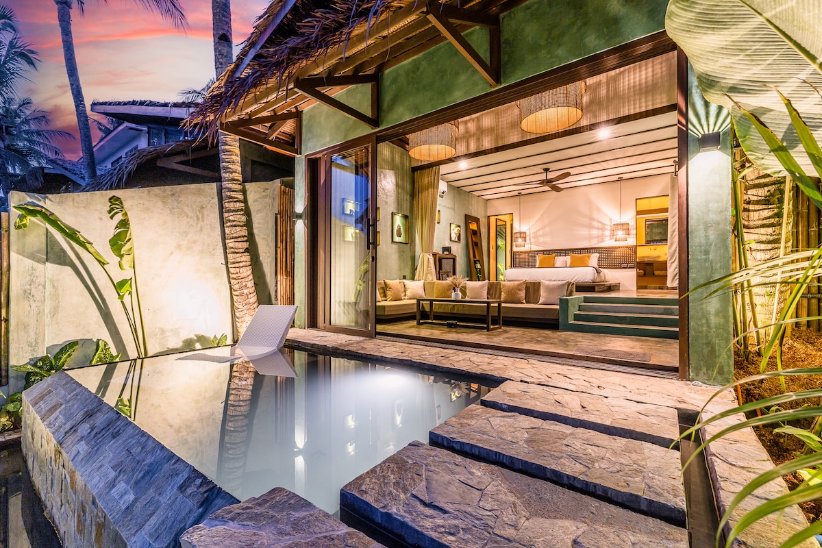 An inviting scene is captured at twilight, showcasing a private infinity pool that merges with the outdoor space. A spacious living area with a king-size bed is visible through expansive glass doors, surrounded by tropical plants and textured stone pathways.