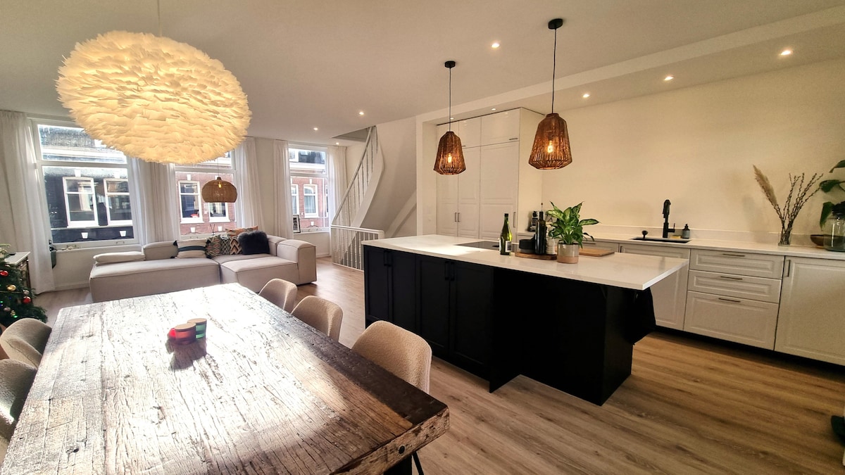 An open-plan living area features a modern kitchen with sleek cabinetry and a large island. A rustic dining table is set beneath a unique chandelier. Bright natural light flows through large windows, creating a welcoming ambiance in the space, which includes a cozy seating area.