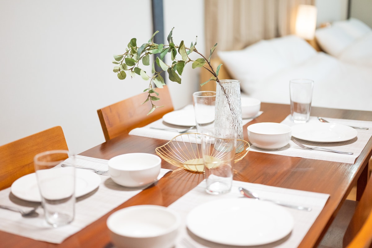 A wooden dining table is set for six, featuring white plates, bowls, and drinking glasses. A decorative vase with greenery adds a natural touch to the arrangement. Soft lighting from the background enhances the inviting ambiance of the space.