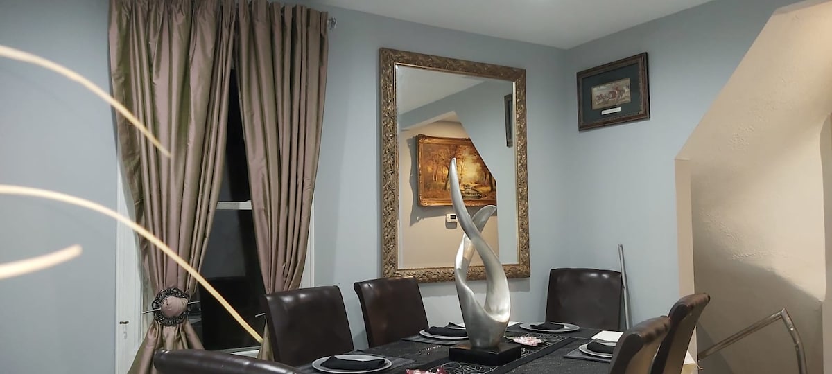 A spacious dining area features a large table surrounded by dark leather chairs. Elegant curtains frame the window, while a decorative mirror enhances the room's depth. A sculptural centerpiece sits on the table, complemented by decorative plates and utensils.