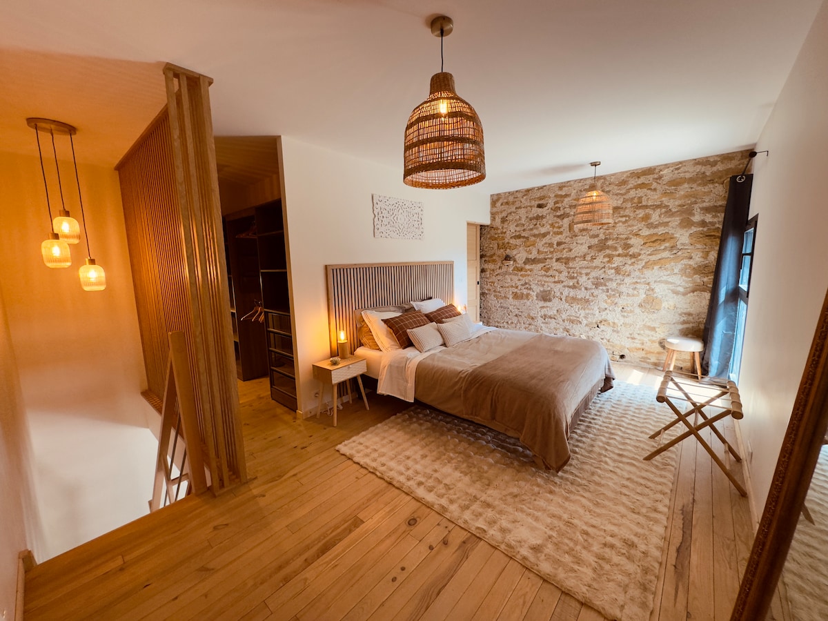 The sleeping area features a spacious bed with neutral-toned bedding against a warm stone wall. Soft lighting is provided by pendant lamps, and a wooden floor enhances the inviting atmosphere. A small side table and a folding chair are positioned nearby for convenience.