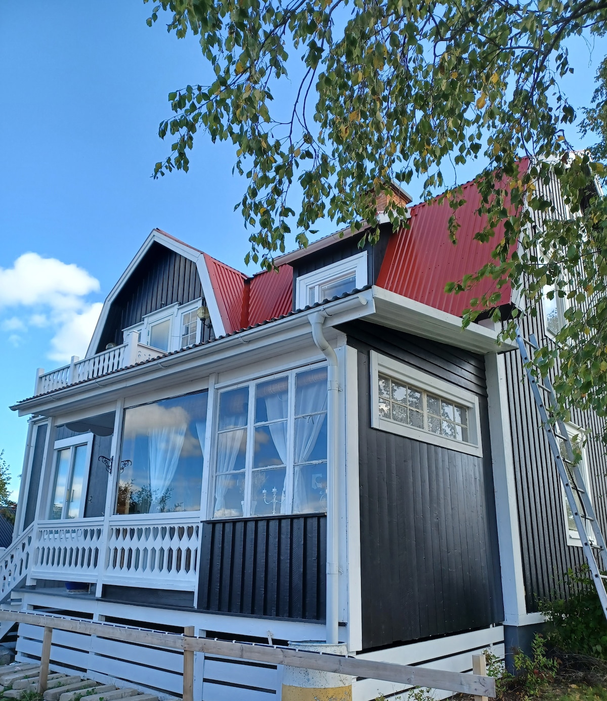 The exterior showcases a charming two-story house with a distinctive red and black panelled facade. A balcony is visible on the upper level, framed by large windows that allow natural light into the interior. Surrounding greenery adds to the house's character.