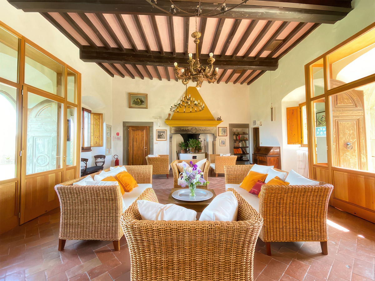 A communal lounge area features comfortable rattan seating arranged around a central table. Bright cushions add subtle color against the warm tones of the room. High ceilings with wooden beams and windows allow natural light to create an open and airy atmosphere.