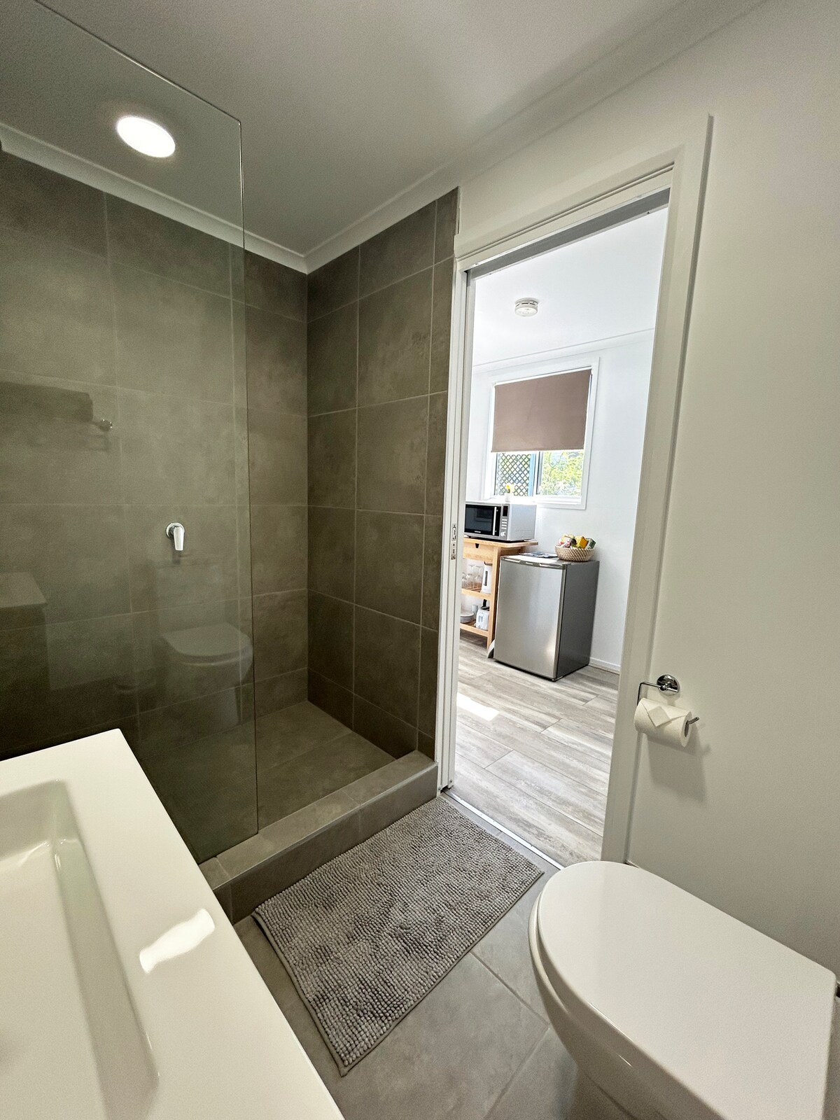 A modern bathroom showcases grey tiled walls and a glass shower enclosure. A sleek sink with a spacious countertop is visible. A door leads to an adjoining room, where a bar fridge and microwave are positioned, contributing to the overall functionality of the space.