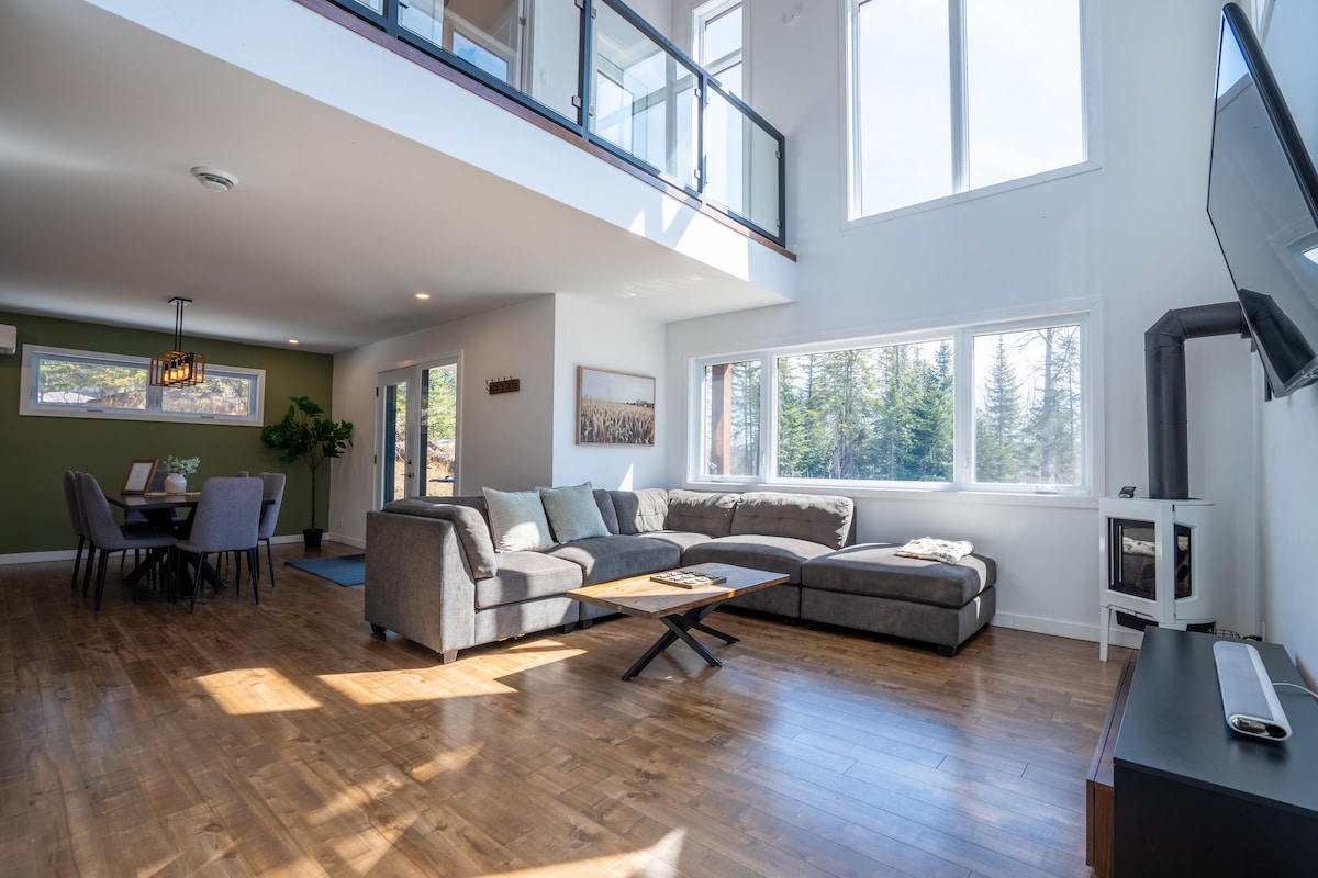The interior living space features large windows allowing natural light to fill the area. A spacious sectional sofa is positioned beside a wooden coffee table. The open floor plan connects the living area to a dining space, where a table is set for six. A fire stove adds warmth to the setting.