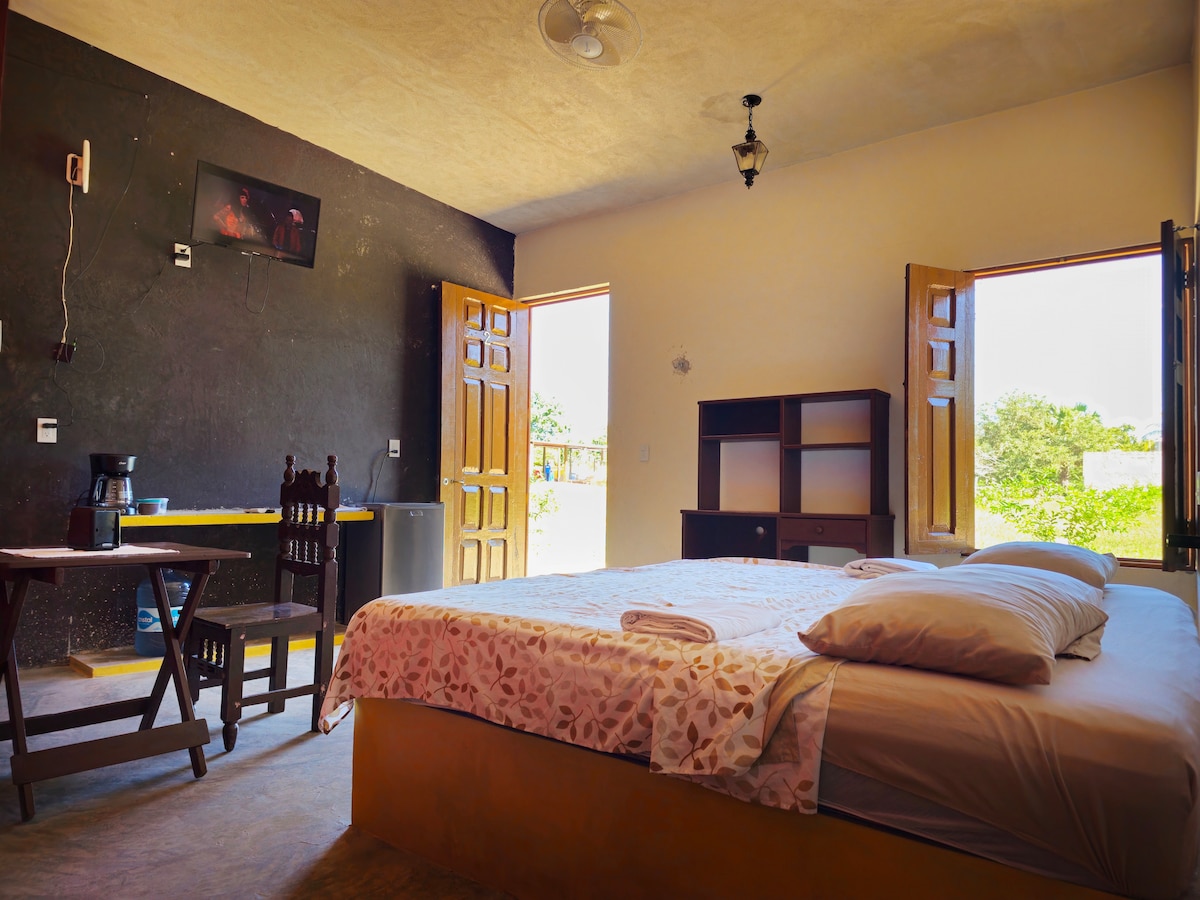 A cozy sleeping area features a spacious bed with light bedding and decorative pillows. A small dining table and chairs are positioned nearby, with a flat-screen TV mounted on the wall. Natural light enters through two open doors, revealing greenery outside.