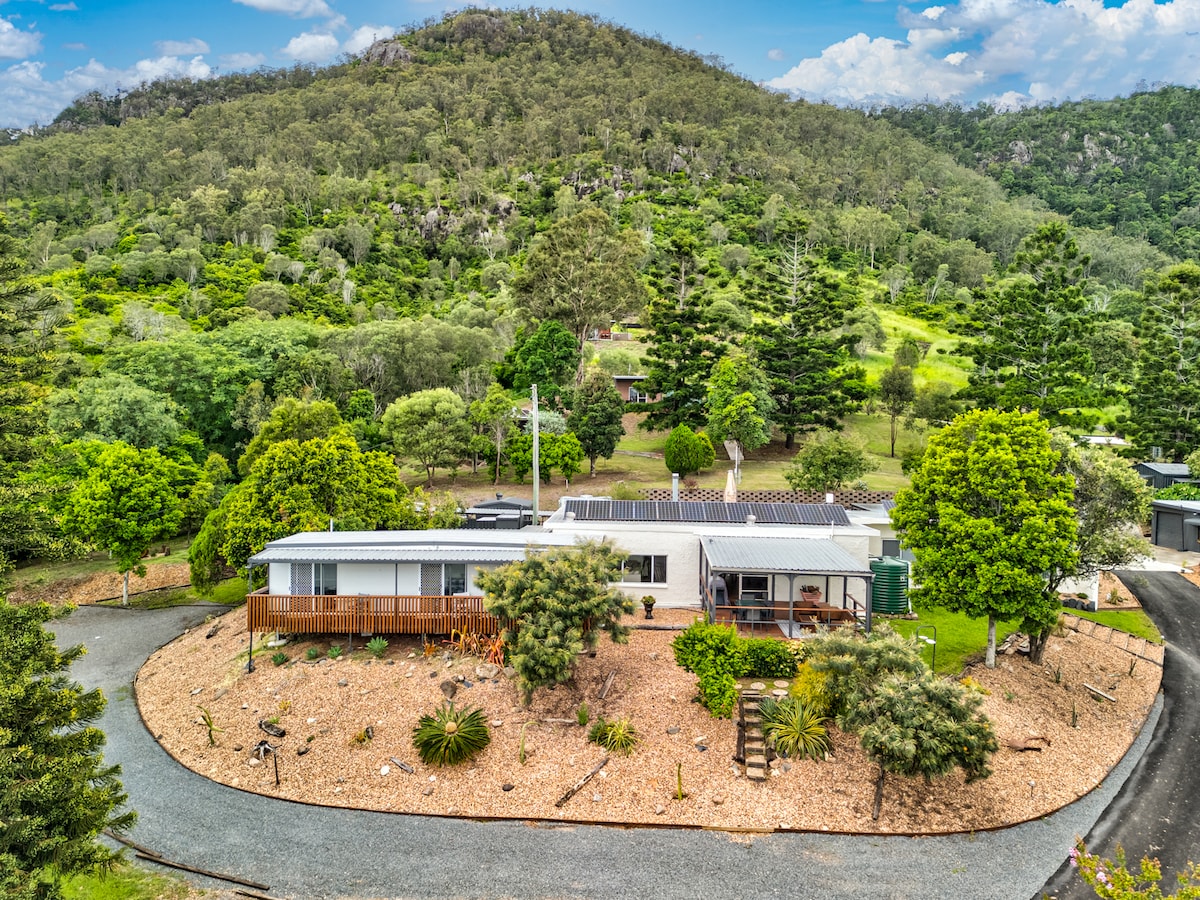 An aerial view of the main house at The Archer is captured, showcasing its modern design surrounded by lush greenery and landscaped gardens. The winding driveway leads to the entrance, while rolling hillside views and trees add to the serene natural setting.