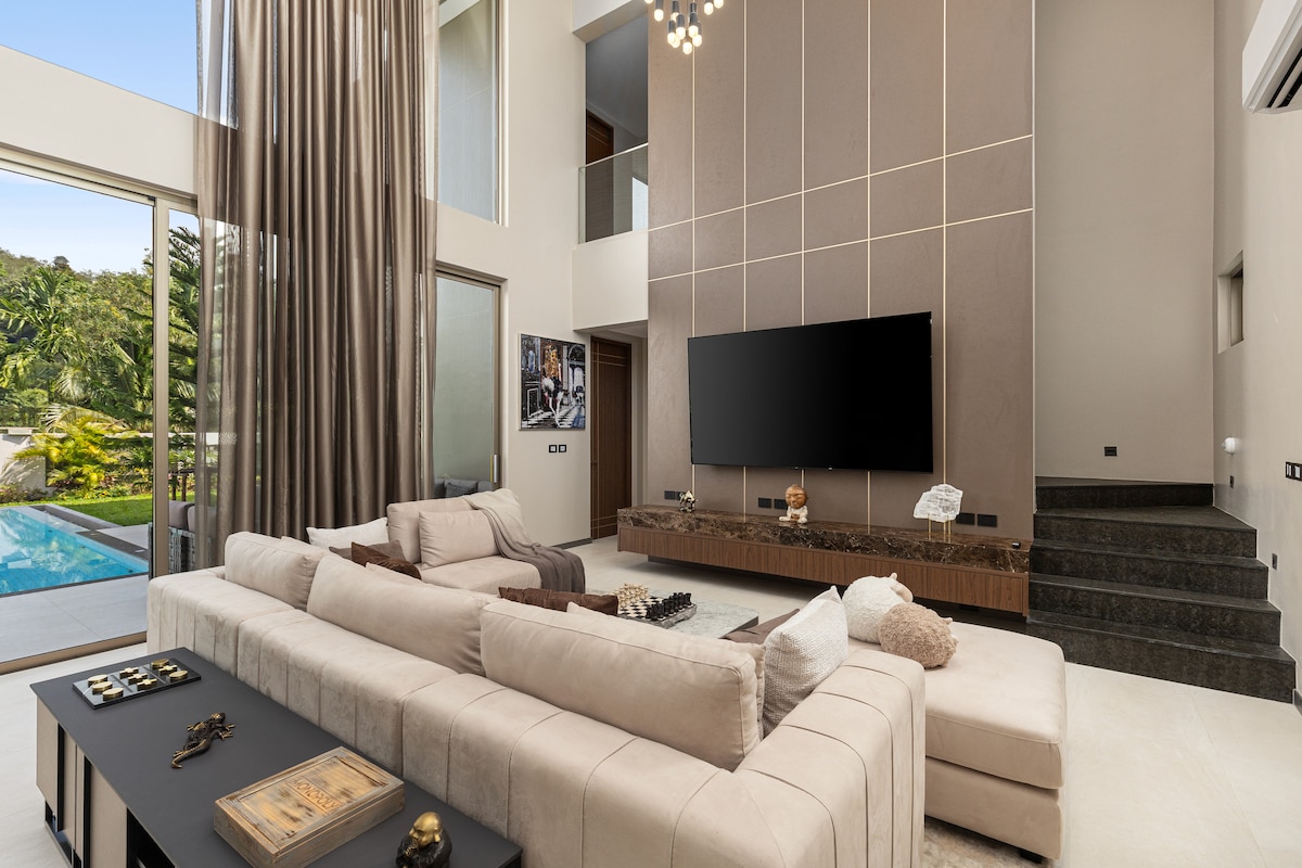 A spacious living area is showcased with a modern sectional sofa and a large television mounted on the wall. Natural light pours in from expansive glass doors leading to the outdoor pool area. The elegant design features soft neutral tones and decorative elements around the room.