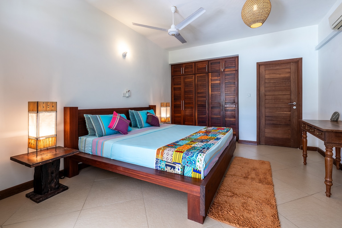 A spacious bedroom features a large bed adorned with colorful decorative pillows. Two wooden bedside lamps provide soft lighting, and a stylish wooden cabinet with multiple doors offers storage. A textured area rug complements the tiled floor.