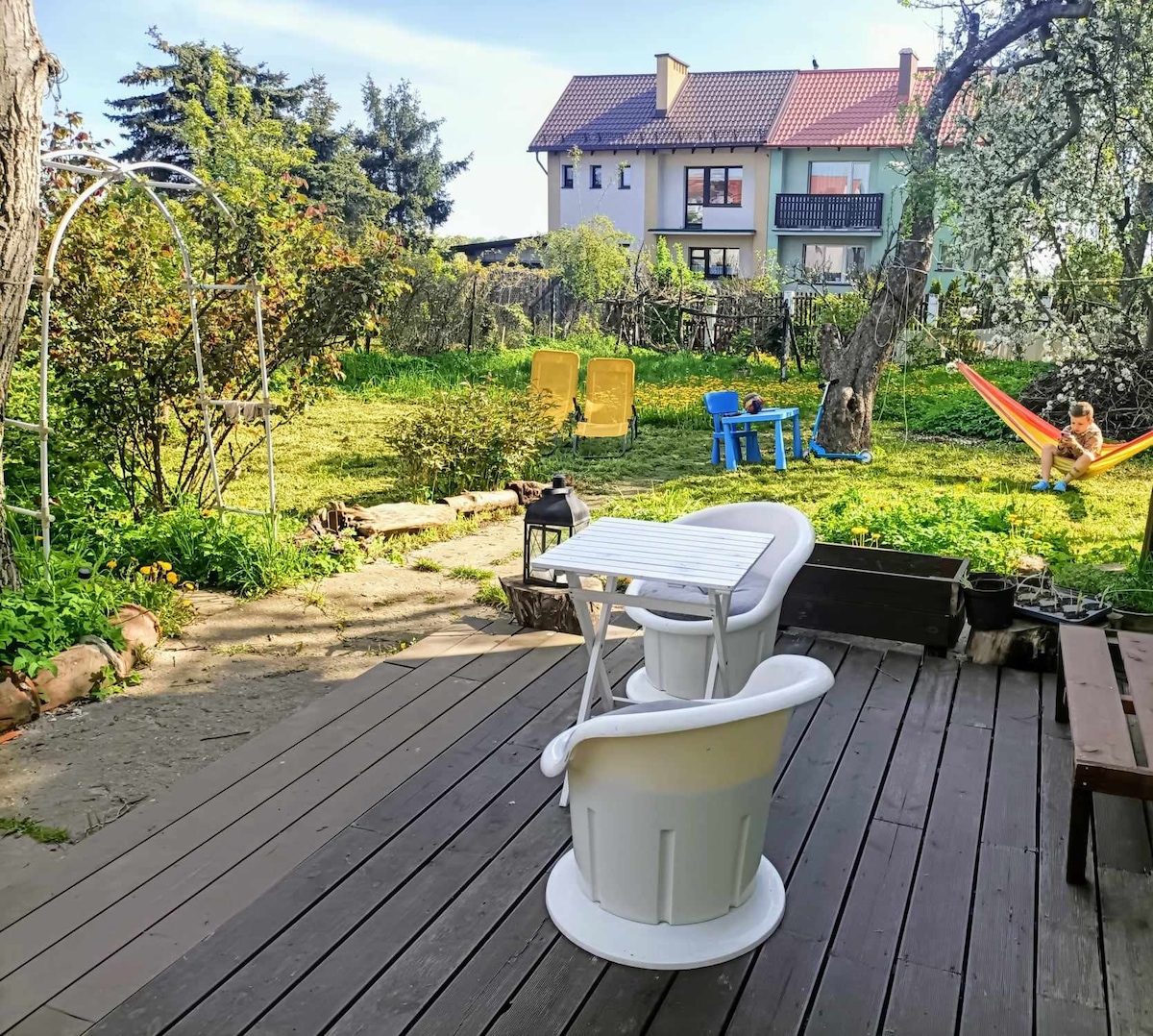 A spacious garden area is visible, featuring a wooden deck with two white chairs and a small table. Vibrant greenery fills the background, with a gazebo and various seating options. A hammock and solitude invite relaxation amidst the surrounding nature.