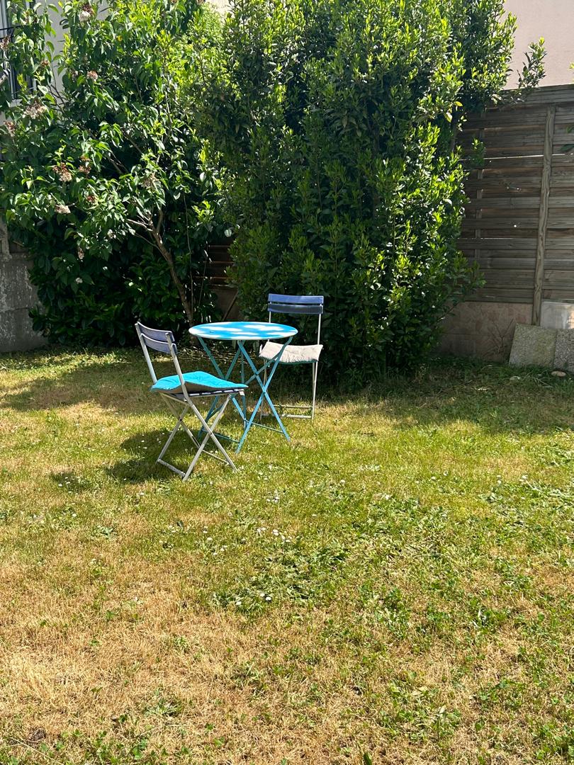 A grassy garden area features a small round table surrounded by two chairs. Lush greenery from nearby trees provides a natural backdrop, enhancing the privacy of the outdoor space.