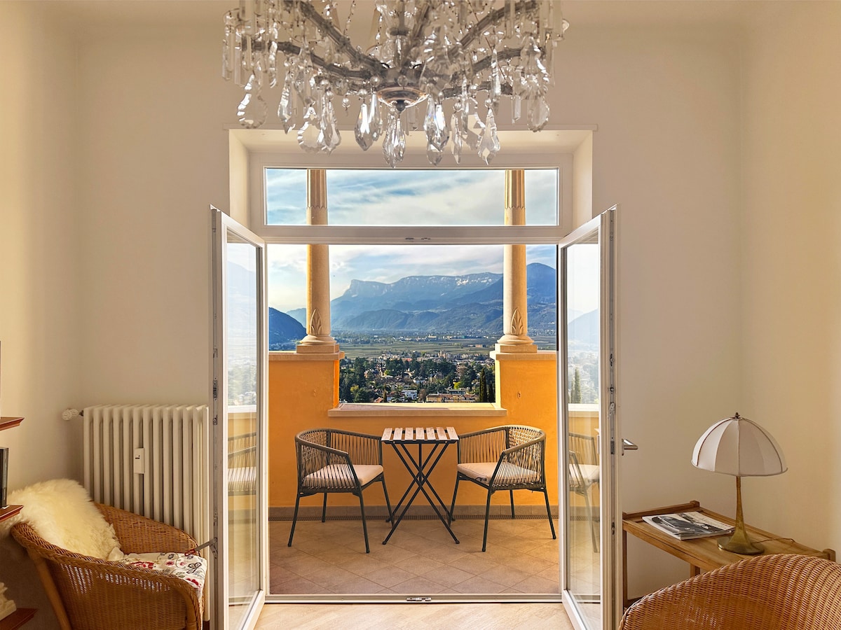 A bright living space features large open doors leading to a balcony. Two outdoor chairs and a small table are positioned to overlook the scenic view of mountains and valleys. A stylish chandelier hangs from the ceiling, adding elegance to the room.