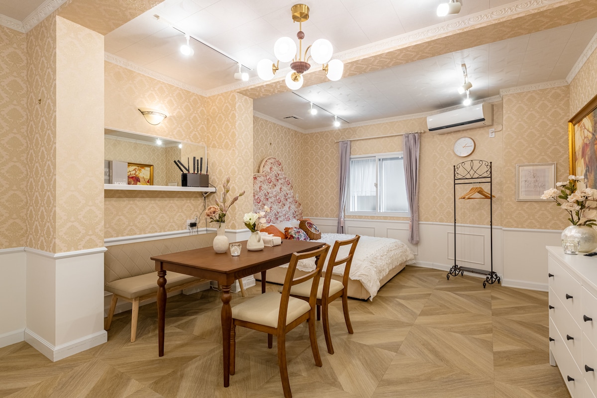An inviting living space features a dining table with wooden surfaces and two chairs. A cozy sleeping area is visible with a bed adorned in white linens. Soft lighting is provided by a central chandelier and wall lamps, creating a warm atmosphere. A decorative mirror and wall accents enhance the room's aesthetic.