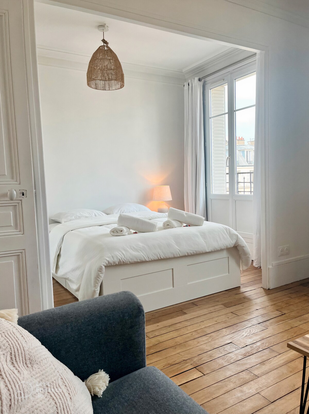 A bright sleeping area is visible, featuring a spacious bed with white linens and four pillows. Natural light floods the room through a large window. A soft lamp provides additional illumination, and warm wooden flooring complements the inviting decor.