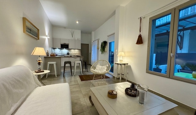 A modern living area is featured with neutral-toned walls and natural light streaming through large windows. A cozy sofa and a decorative side table are positioned next to a dining area, which includes a kitchen with contemporary cabinetry and essential appliances.