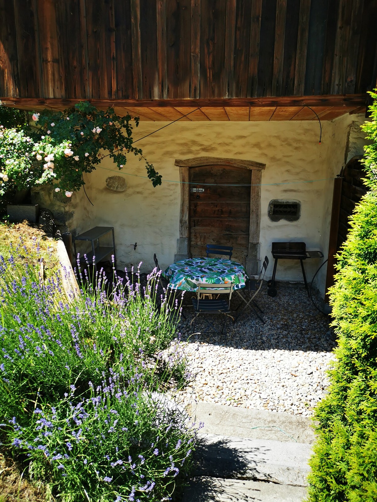 A private outdoor space is presented with a small table and chairs set among lavender plants. A stone path leads towards a rustic wooden door, framed by climbing greenery, creating a tranquil entrance to the chalet.