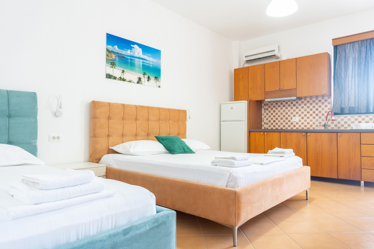 A spacious room features two well-appointed beds with premium linens and a decorative pillow. A kitchenette with wooden cabinetry is visible, alongside a seating area. Natural light enters through a window with dark curtains, and beach-themed art adorns the wall.