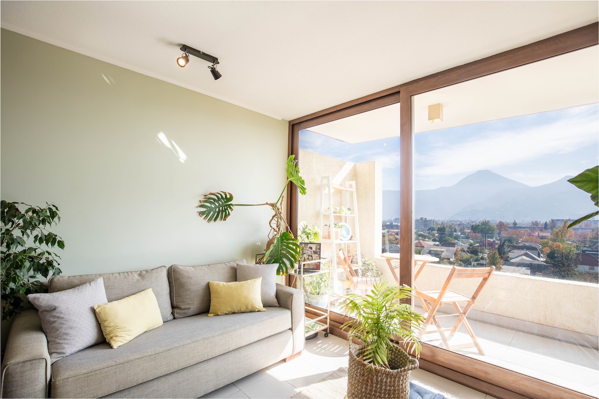 A light-filled living area features a neutral-toned sofa with yellow accents. Large glass doors open to an inviting terrace, showcasing a wonderful view of the surroundings. A potted plant and a small wooden table complement the space, enhancing the connection to the outdoors.