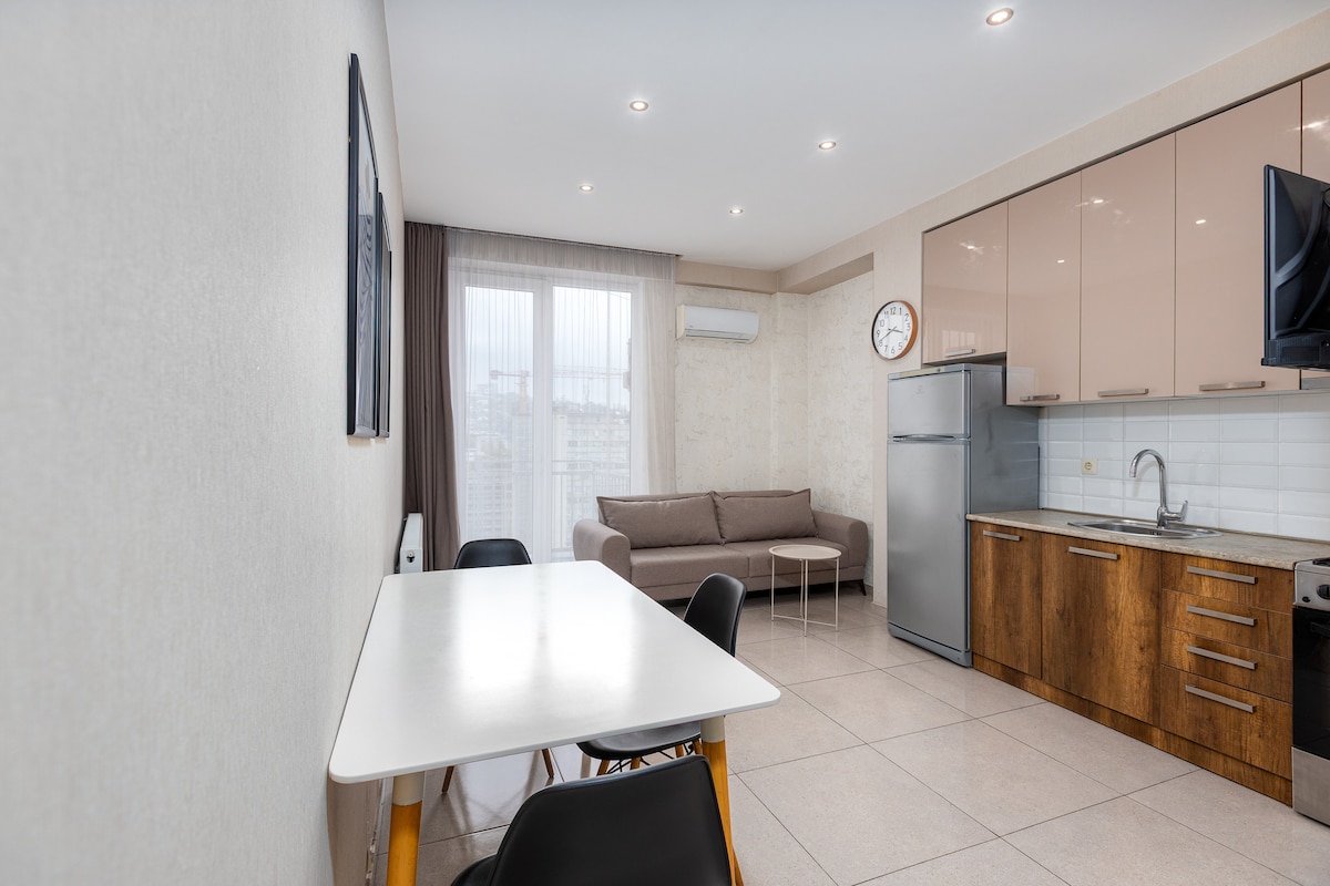 The joint kitchen and living space features a modern design with sleek cabinetry and a stainless steel refrigerator. A small dining table with black chairs provides a dining area, while a comfortable sofa is positioned nearby. Large windows allow natural light to fill the room.