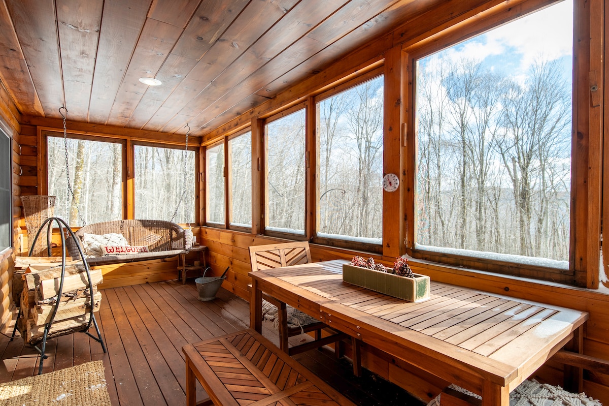 A screened porch is lined with large windows, providing views of surrounding trees. Wooden flooring complements a dining table set for four and a cozy seating area with a hammock chair. Natural light floods the space, creating a serene environment for relaxation.