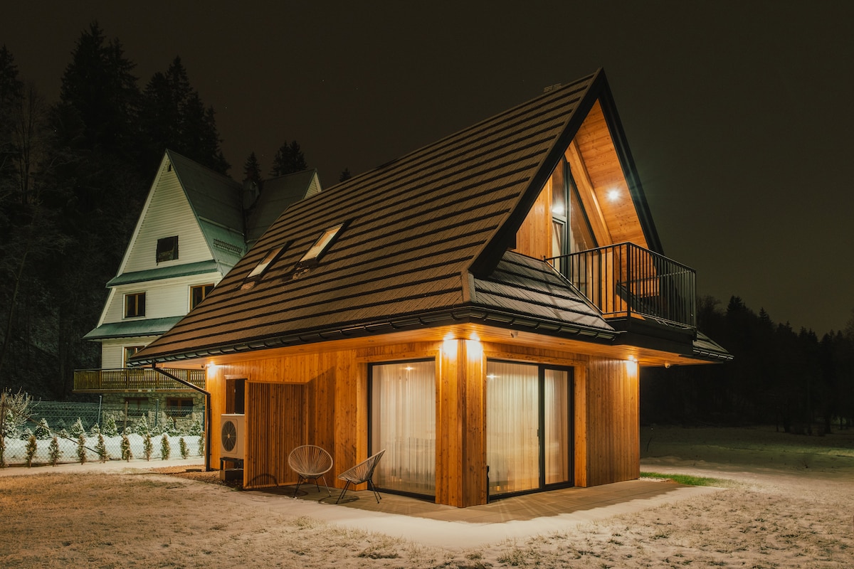 A modern chalet is illuminated at night, featuring a sloped roof and wooden exterior. Large windows provide a view into the cozy interior, and a balcony extends from the second floor. Surrounding snow and vegetation create a serene atmosphere.