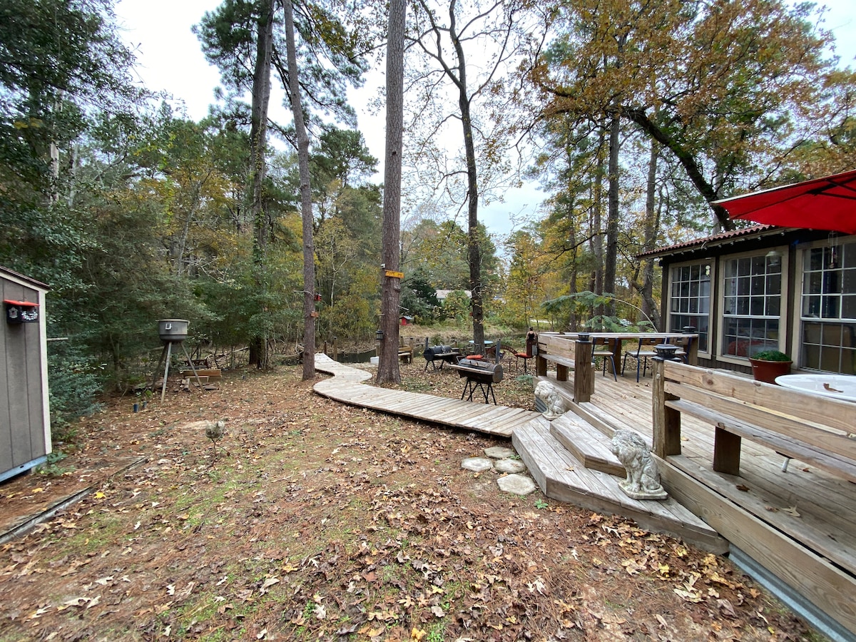 A wooden walkway meanders through a natural setting, leading to a spacious deck area equipped with outdoor seating. The surrounding trees provide shade, and fallen leaves cover the ground, indicating the seasonal change. A hot tub is visible near the deck for relaxation.