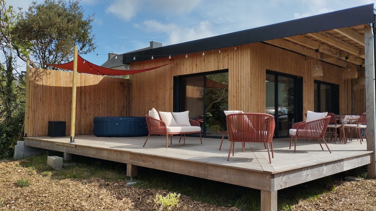 A spacious outdoor terrace showcases comfortable seating arrangements with modern, red-accented chairs. A hot tub is visible in the corner, framed by wooden slats and soft lighting, while a vibrant awning provides shade. Lush greenery surrounds the area, creating a serene atmosphere.