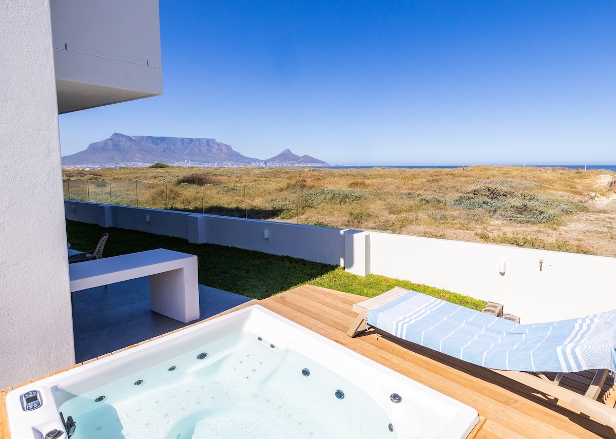 A spacious outdoor area features a private whirlpool alongside a sun lounger. In the background, expansive views of the ocean and Table Mountain can be seen, highlighting the natural beauty of the surroundings.