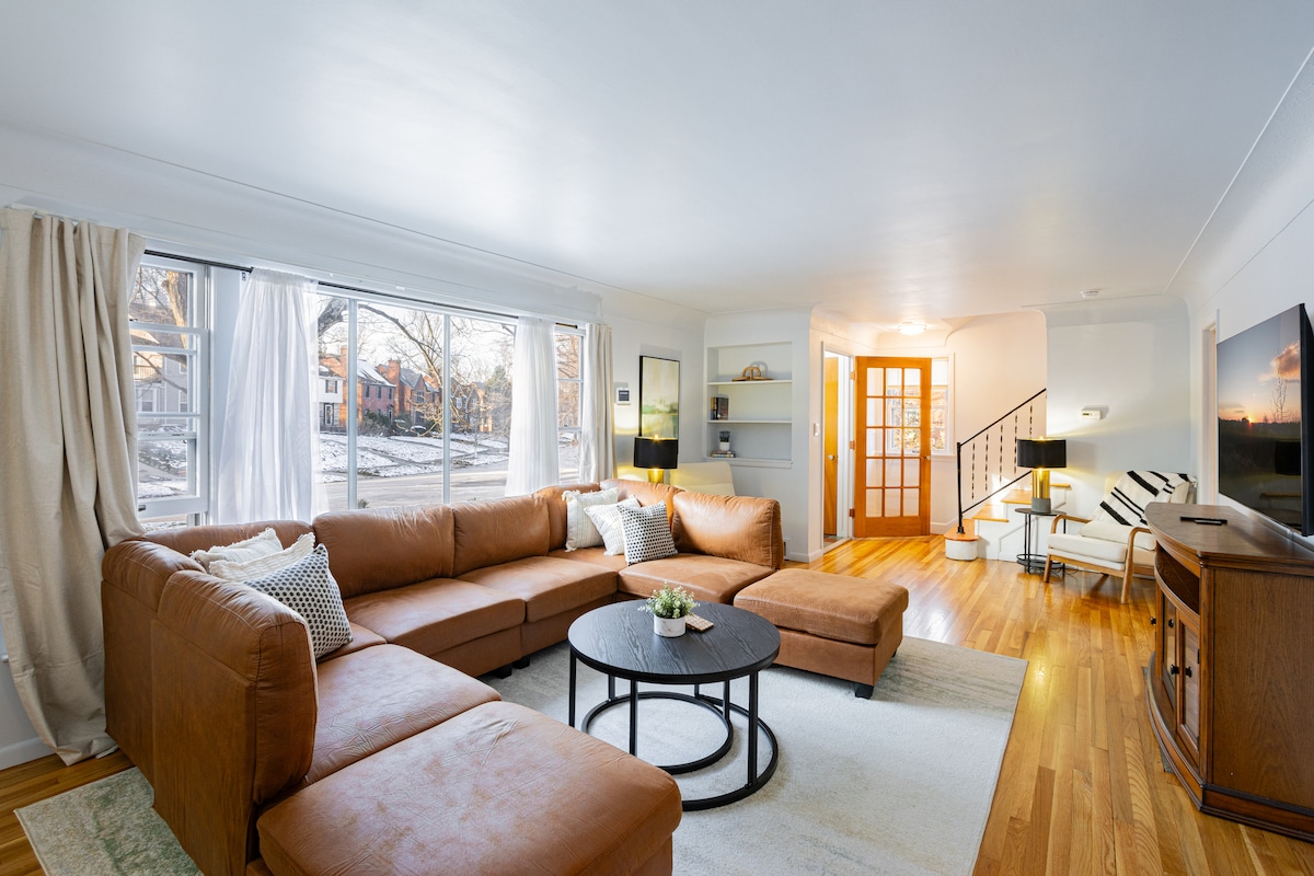 A light-filled living area features a large sectional couch in a warm tan color, paired with a round coffee table. Windows let in natural light, and a side table with a lamp is positioned beside the couch. A television is mounted on a wall opposite the seating.