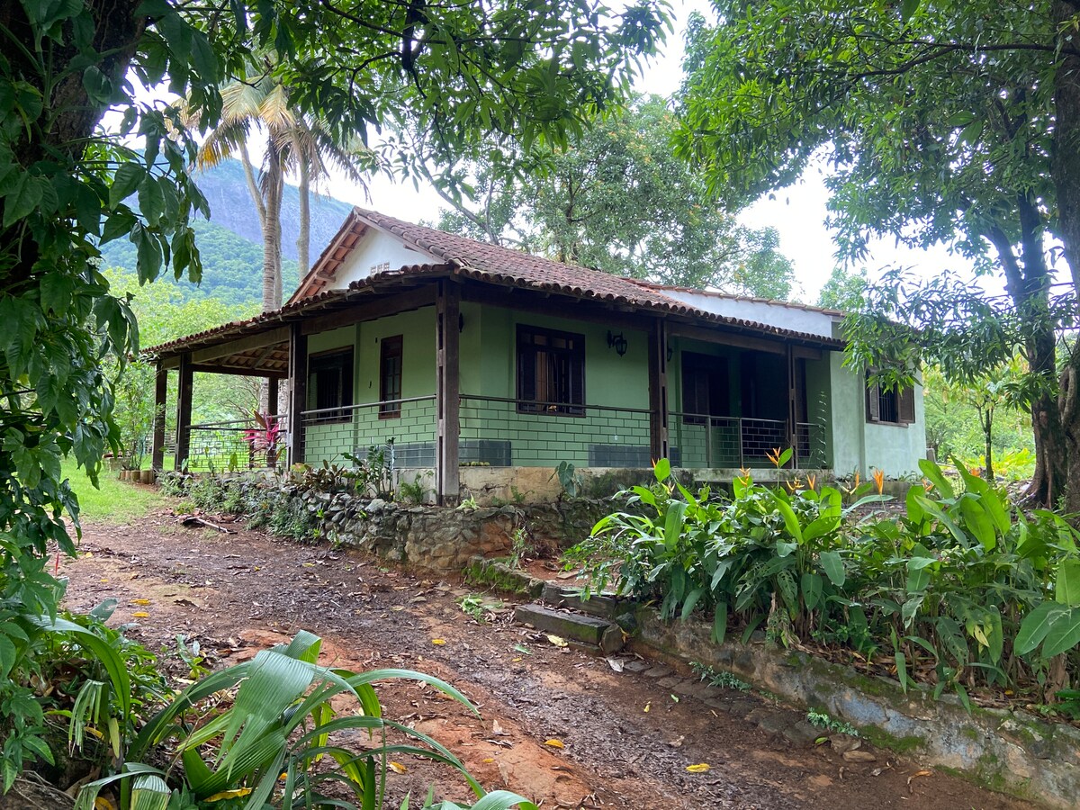 The house is surrounded by lush greenery, featuring a rustic design with a sloped roof and wooden accents. A spacious porch extends along the front, offering views of the surrounding nature. The pathway leading to the house is lined with natural vegetation.