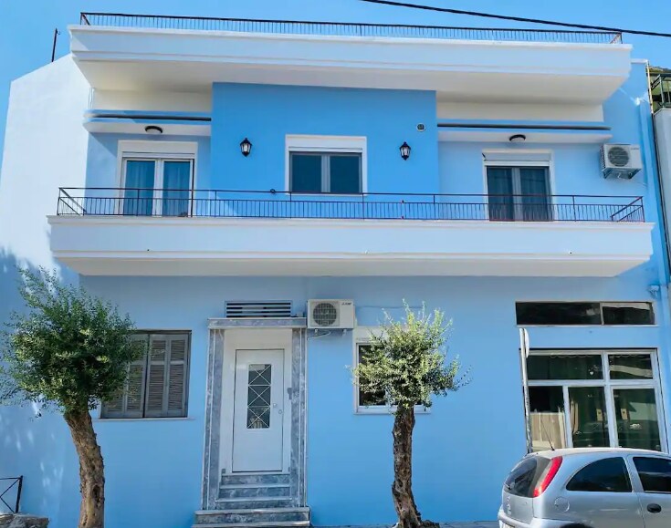 The exterior of a recently renovated blue building features a simple, modern design. Two small trees frame the entrance, which is accented by a marble threshold. Balconies are visible on the upper level, and large windows contribute to a welcoming appearance.