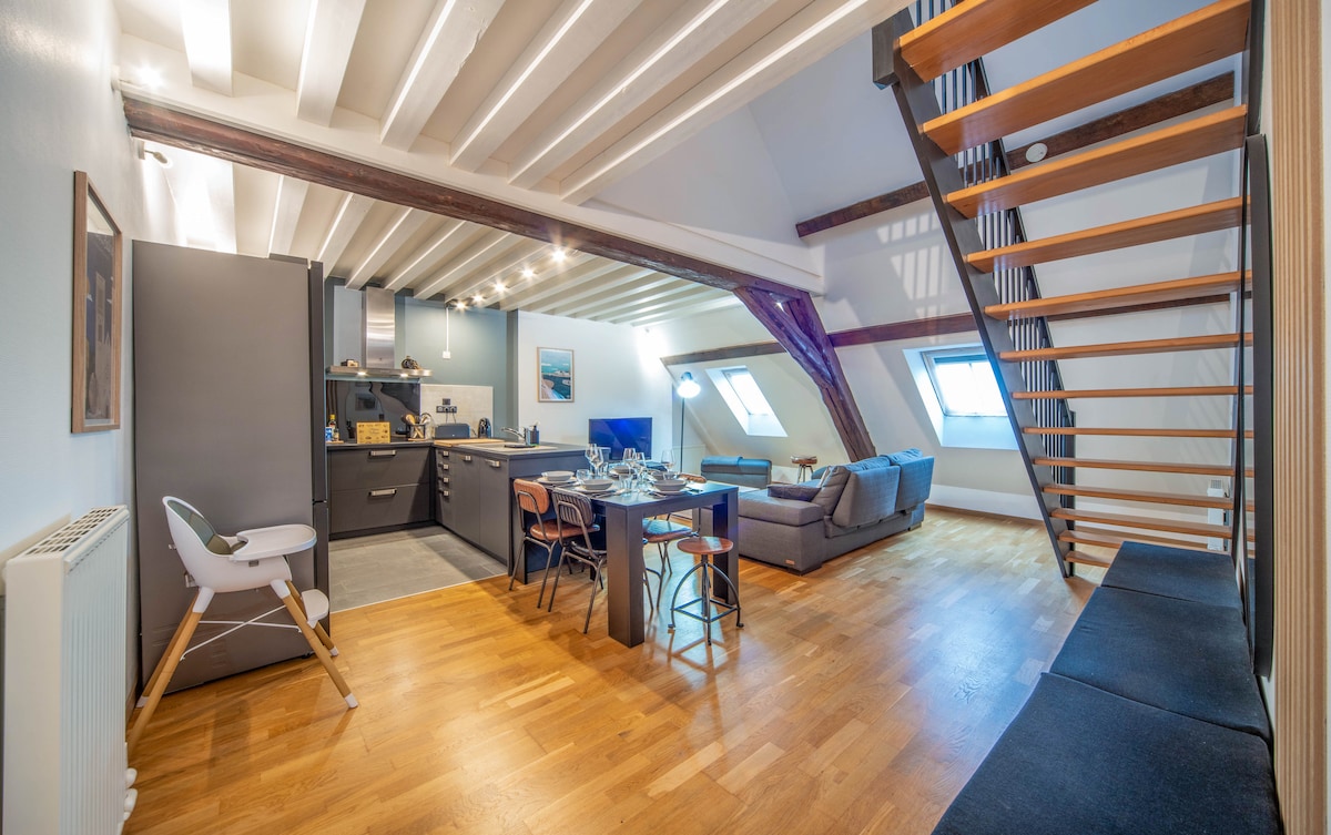An open-plan living area features a well-equipped kitchen with modern appliances, a dining table set for meals, and a comfortable seating area with a large sofa. Natural light floods in through angled windows, while stairs lead to a mezzanine level.