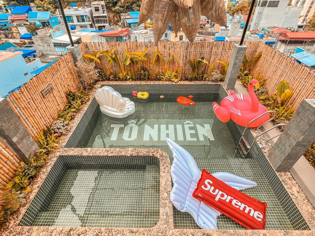 A rectangular pool is surrounded by greenery and bamboo fencing. Colorful pool floats, including a flamingo and shell, are scattered throughout the water. The bottom of the pool displays the name 'TÔ NHIÊN', providing a fun focal point in the tranquil setting.