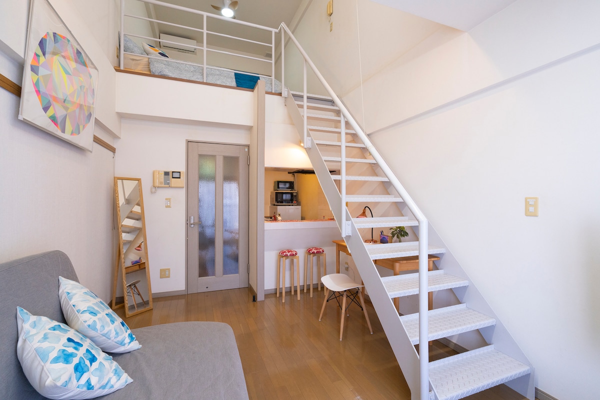 A two-level space features a staircase leading to an upper sleeping area, while a cozy living space includes a gray sofa and a mirror. The kitchen area is visible, offering a practical layout with bar stools and a small dining table.