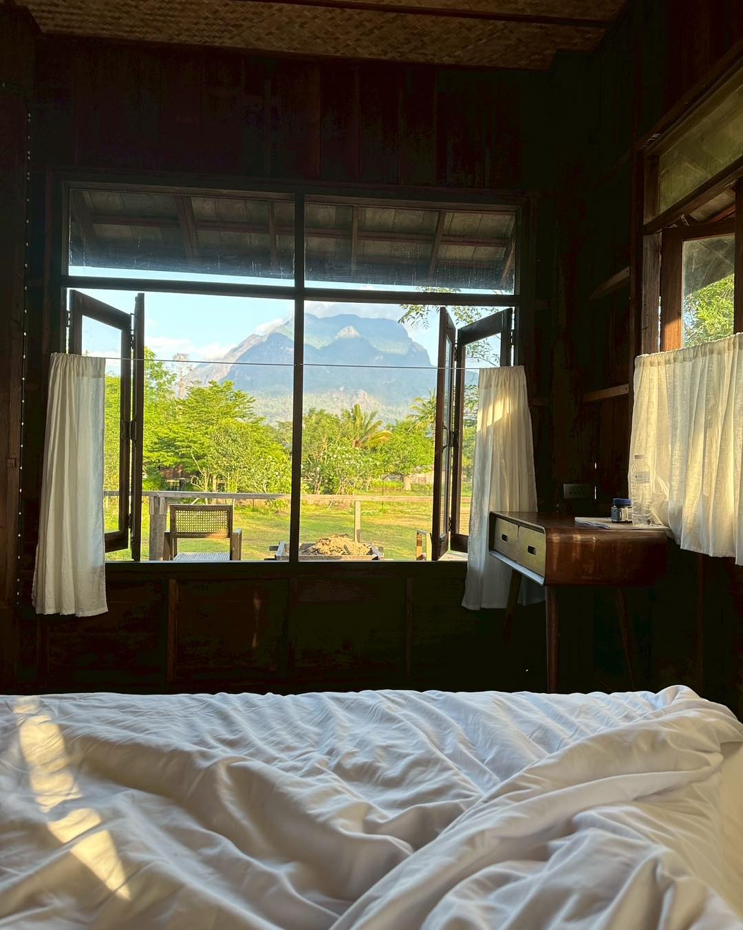 A serene bedroom is featured with a spacious bed draped in soft white linens. Large windows frame a view of lush greenery and majestic mountains, inviting natural light into the room. The wooden elements and rural landscape create a peaceful atmosphere.