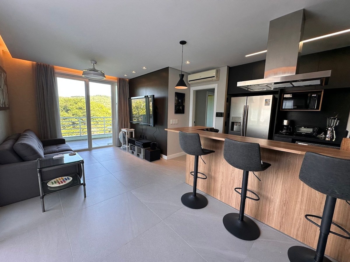An open-concept living area features a sleek kitchen with a modern bar area and high stools. A comfortable seating arrangement is positioned nearby, and large windows provide natural light, revealing views of greenery outside.