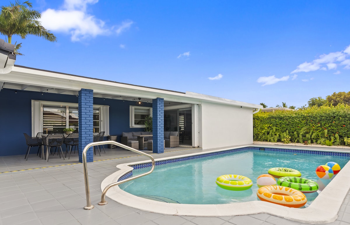 A heated pool with colorful floaties is featured in the foreground, surrounded by a tiled deck. A patio area with seating is visible, complemented by sleek blue and white walls. Lush greenery can be seen in the background, enhancing the outdoor space.