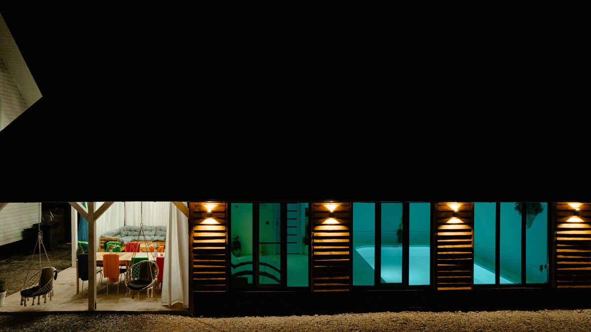 Illuminated windows frame a modern cabin at night, showcasing an inviting indoor dining area through sheer curtains. A tranquil pool gleams in turquoise, contrasted by warm lights enhancing the property's exterior. A cozy outdoor seating area is visible, offering a blend of relaxation and comfort.
