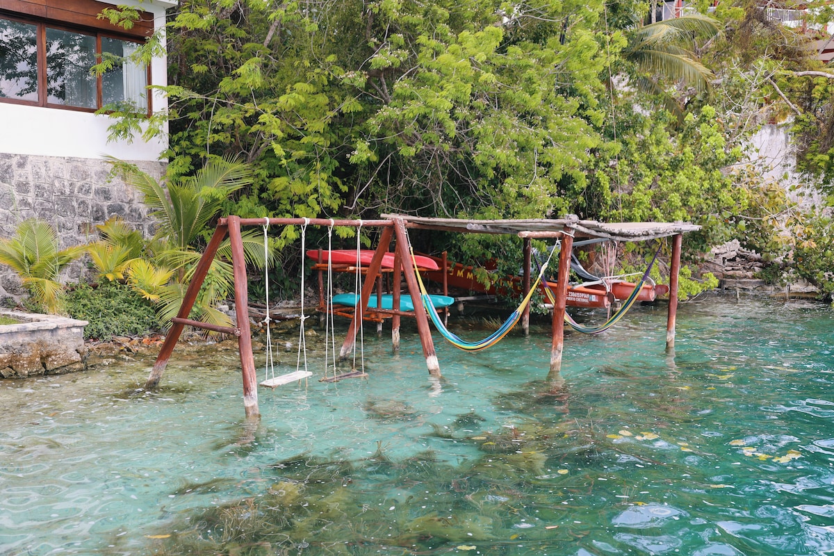 A wooden structure is positioned over clear water, featuring swings and hammocks intertwined with lush greenery. Kayaks are visible nearby, adding to the recreational options available at the lagoon. The serene aquatic environment complements the natural surroundings.