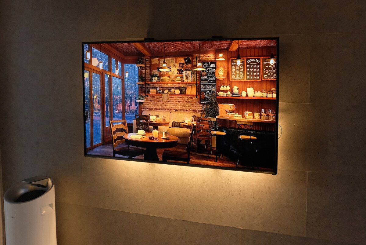 A framed image displays a warm cafe setting featuring wooden tables and chairs. A brick wall enhances the rustic atmosphere, while shelves in the background display various food items. Natural light filters in through large windows, creating an inviting and cozy ambiance.